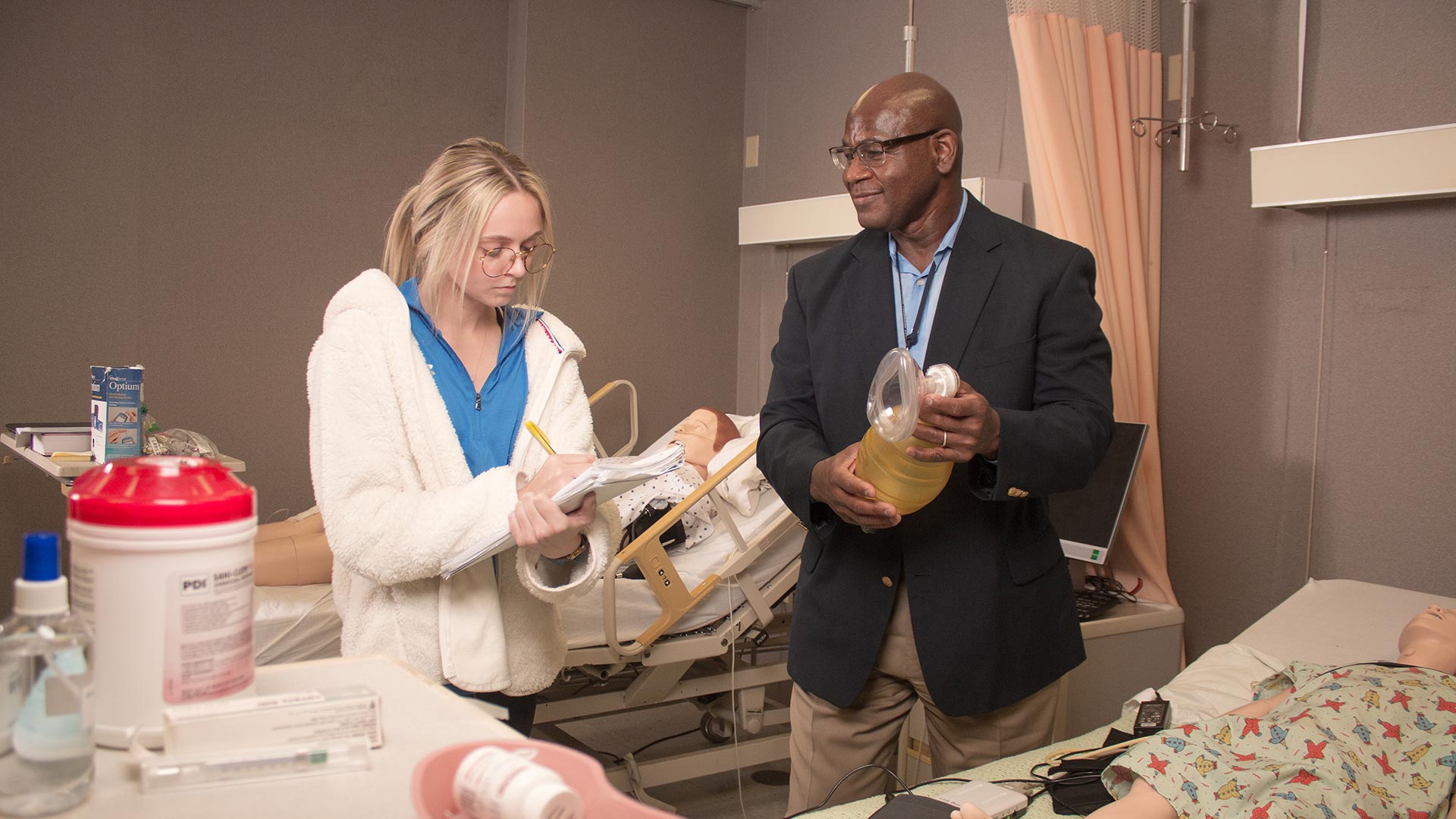 Female student taking notes while Male professor demonstrates nursing things