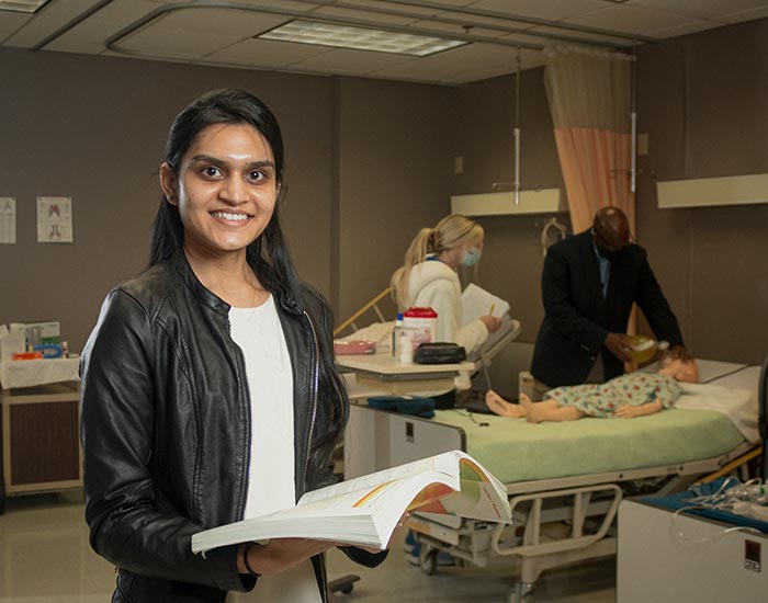 female student holding book while student/teacher check on kid in hospital bed in background